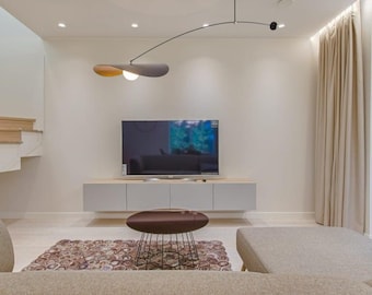 May include: A modern living room featuring a large flat-screen TV mounted above a gray cabinet. A brown and black coffee table sits on a patterned rug. A unique, curved light fixture hangs from the ceiling, and beige curtains frame the window.