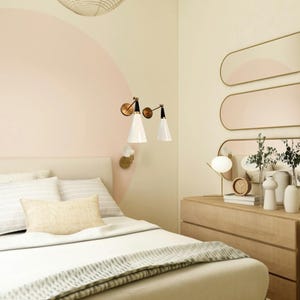 May include: A bedroom scene with a bed, dresser, and wall decor. The bed has white bedding and a light-colored headboard. Two white cone-shaped lamps are mounted on the wall. A dresser with a mirror and decorative items is on the right.