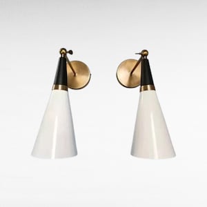 May include: Two mid-century modern wall sconces. Each sconce features a white, cone-shaped shade, a black top section, and a brass-colored mounting plate and arm. The sconces are designed to be mounted on a wall and provide directional lighting.