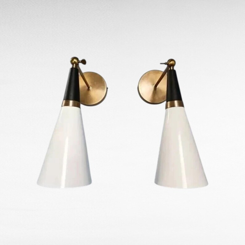May include: Two mid-century modern wall sconces. Each sconce features a white, cone-shaped shade, a black top section, and a brass-colored mounting plate and arm. The sconces are designed to be mounted on a wall and provide directional lighting.