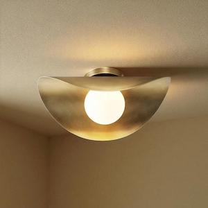 Op de afbeelding: Een messingkleurige plafondlamp met een halvemaanvormige kap en een bolvormige gloeilamp. De lamp is gemonteerd op een neutraal gekleurd plafond. De kap heeft een gebogen, organische vorm en de gloeilamp straalt een warme gloed uit.