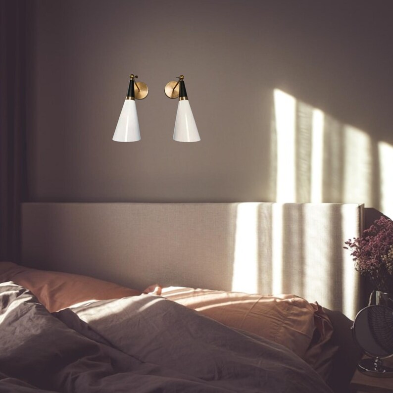 May include: Two modern wall sconces with white cone-shaped shades, black accents, and brass-colored circular backplates. The sconces are mounted on a neutral-colored wall above a bed with peach and gray bedding. The room is lit by natural light.