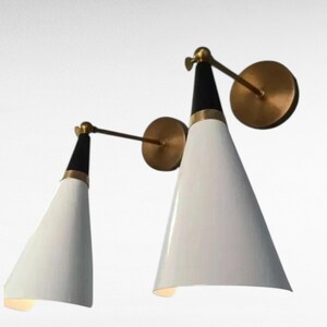 May include: Two modern wall sconces with white conical shades, black tops, and brass accents. The sconces have a pivoting arm and a circular wall mount. The design is minimalist and stylish, suitable for interior lighting.