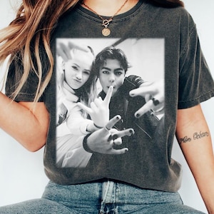 May include: Dark grey t-shirt with a black and white photo of two people making hand gestures. The photo is centered on the shirt. The shirt has a crew neck and short sleeves. The model is wearing a gold necklace and blue jeans.