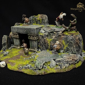 May include: A detailed miniature wargaming terrain piece. It features a stone structure with an archway, rocky terrain, and miniature figures. The terrain is detailed with green moss and a stone path. Dice are visible.