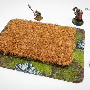 May include: A rectangular wargaming terrain piece with a brown, textured center and green edges. Miniature figures and dice are placed around the terrain. The terrain piece is designed for tabletop gaming.