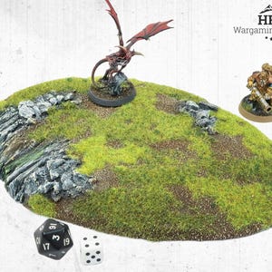 May include: A miniature wargaming terrain piece with a grassy hill and rocky cliffs. A red dragon figure and a soldier figure are placed on the terrain. Dice are also included. The terrain is labelled "HRC Wargaming Terrain."