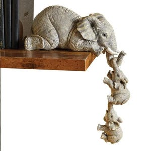 Puede incluir: Una figura de elefante gris que descansa sobre una estantería de madera, con tres elefantes más pequeños colgando de su trompa. Los elefantes tienen piel texturizada y son un artículo decorativo.