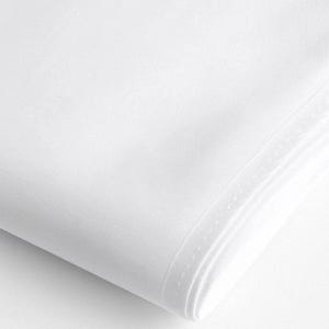 Puede incluir: Primer plano de una tela blanca doblada. La tela parece lisa y de tejido apretado, con un ligero brillo. Los bordes están cuidadosamente acabados con una costura sutil. La impresión general es de un textil limpio y nítido.