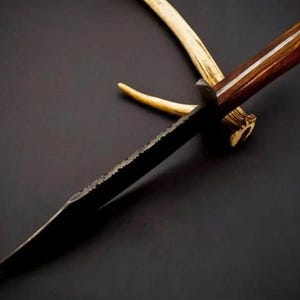 May include: A fixed-blade knife with a dark, textured metal blade and a polished brown wooden handle. The knife is positioned over a light-colored, curved antler piece. The blade features a jagged, uneven spine and a sharp, pointed tip.