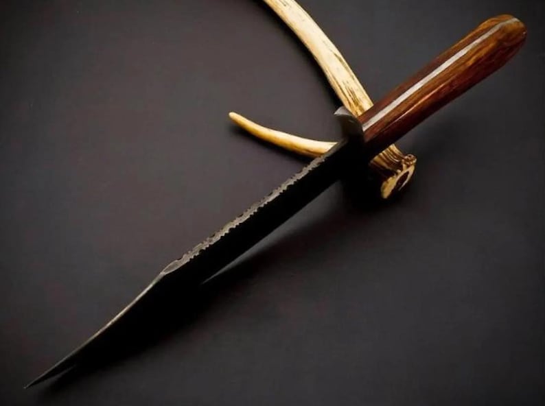 May include: A fixed-blade knife with a dark, textured metal blade and a polished brown wooden handle. The knife is positioned over a light-colored, curved antler piece. The blade features a jagged, uneven spine and a sharp, pointed tip.