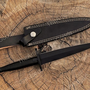 May include: A black dagger with a textured handle and a matching black sheath. The sheath is made of leather and has white stitching around the edges. The dagger and sheath are displayed on a wooden surface.