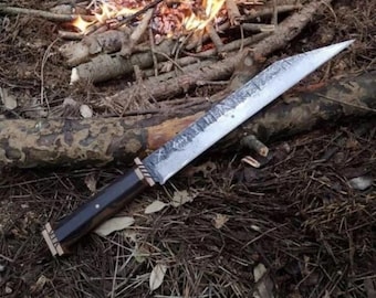 May include: A fixed-blade knife with a long, pointed steel blade featuring a textured, hammered finish and etched markings. The handle is made of dark, polished wood with metallic accents at the bolster and pommel, secured by a single visible pin.