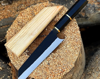 May include: A black and silver chef's knife with a wooden sheath rests on a wood stump covered in wood shavings. The knife has a black handle with gold accents and a silver blade. The wooden sheath is light brown.