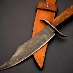 May include: A fixed-blade knife with a dark, mottled steel blade, a wooden handle secured with small metal pins, and a curved metal crossguard. The knife is accompanied by a tan leather sheath featuring decorative stitching and a metal rivet.