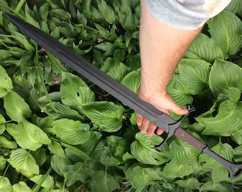 May include: A black sword with a brown handle and a distinctive guard is held against a backdrop of green foliage. The sword's blade is long and sharp, with a dark finish. The handle appears to be wrapped in brown leather.