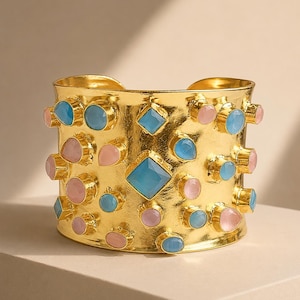 May include: A wide gold-tone cuff bracelet featuring a textured metallic surface adorned with an assortment of scattered, bezel-set gemstones in shades of light blue and soft pink, including round, pear-shaped, and a central square-cut stone.