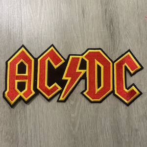 May include: Embroidered patch featuring the AC/DC logo in red and yellow on a black background. The patch has a lightning bolt in the center of the logo. The patch is rectangular and has a yellow border.