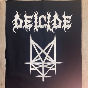 May include: Black square patch with the word "DEICIDE" in white, stylized, flame-like lettering. Below the text is a white, inverted pentagram design. The patch is likely for a jacket or bag.