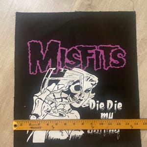 May include: Black and white patch with a pink "Misfits" logo. A woman with a skull face is holding a glass of wine. The text "Die Die Mu" is below the woman.