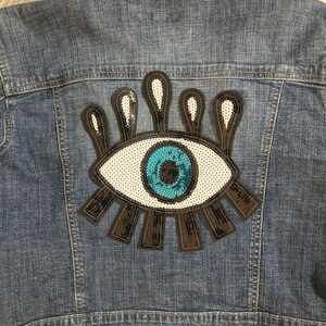 May include: A blue denim jacket with a large, colorful eye patch sewn on the back. The eye is made of sequins and has a white iris, a blue pupil, and black eyelashes.