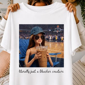Peut inclure: Un t-shirt blanc avec une impression graphique représentant une poupée portant une casquette en jean bleu et des lunettes de soleil sombres, tenant un grand café glacé. La poupée est assise sur une chaise pliante bleue dans un stade de basket. Le texte indique, literally just a bleacher creature.