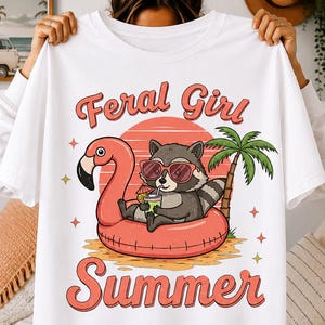 Peut inclure: T-shirt blanc avec un motif d'un raton laveur portant des lunettes de soleil, se relaxant sur un flotteur flamant rose. Le texte "Feral Girl Summer" est affiché avec un coucher de soleil et un palmier.