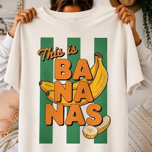 May include: An off-white short-sleeve t-shirt featuring a graphic print of yellow bananas and the text "This is BA NA NAS" in bold orange letters. The design includes three vertical green stripes behind the fruit and a sliced banana piece.
