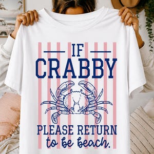 Peut inclure: T-shirt blanc avec un motif de crabe et le texte "IF CRABBY PLEASE RETURN to be beach." Le motif présente une illustration de crabe bleu et des rayures verticales roses et bleues.