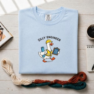 Peut inclure: T-shirt bleu clair avec l'inscription "SILLY ENGINEER" et un dessin d'oie portant un casque jaune et un gilet de sécurité. L'oie tient un plan et un presse-papiers avec un symbole d'engrenage. Une tasse de café, un carnet et un livre ouvert sont également visibles.