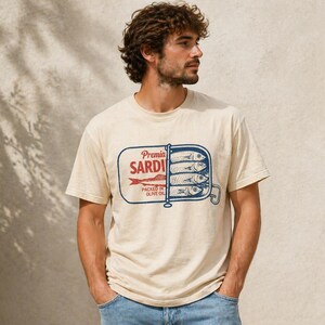May include: A cream-colored short-sleeve t-shirt featuring a graphic print of an open sardine tin. The design includes the text Premia SARDINES PACKED IN OLIVE OIL in red and blue ink, depicting four fish stacked inside a rectangular metal container.