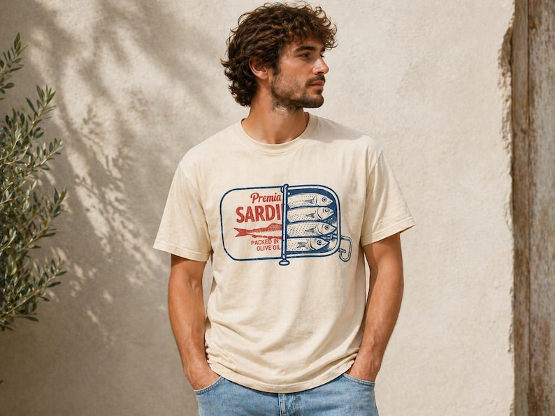 May include: A cream-colored short-sleeve t-shirt featuring a graphic print of an open sardine tin. The design includes the text Premia SARDINES PACKED IN OLIVE OIL in red and blue ink, depicting four fish stacked inside a rectangular metal container.