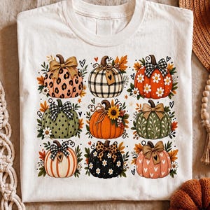 Peut inclure: Un t-shirt blanc orné d'une impression graphique représentant neuf citrouilles disposées en grille. Chaque citrouille présente des motifs uniques comme des taches de léopard, des carreaux, des pois et des fleurs, agrémentés de nœuds et de feuilles.