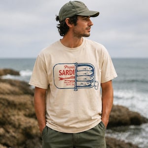 May include: A beige short-sleeve t-shirt featuring a graphic of an open sardine tin with the text Premia SARDINES PACKED IN OLIVE OIL. The design includes a red fish illustration and blue line art of sardines stacked inside the rectangular tin container.