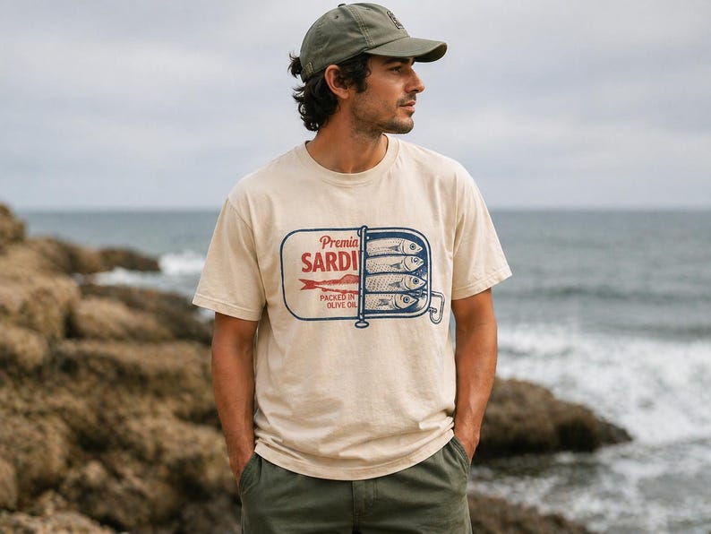 May include: A beige short-sleeve t-shirt featuring a graphic of an open sardine tin with the text Premia SARDINES PACKED IN OLIVE OIL. The design includes a red fish illustration and blue line art of sardines stacked inside the rectangular tin container.