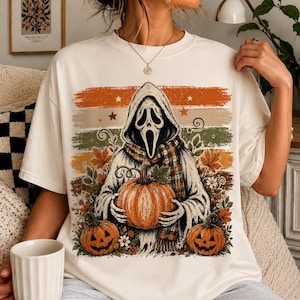 May include: An off-white short-sleeve t-shirt featuring a graphic of a hooded ghost figure wearing a plaid scarf and holding a pumpkin. The design includes two additional jack-o'-lanterns, autumn leaves, and horizontal stripes in orange, tan, and green.