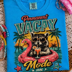 Könnte beinhalten: Hellblaues T-Shirt mit der Aufschrift "Permanent Vacay Mode On" und einer Grafik eines Waschbären mit Sonnenbrille auf einem rosa Flamingo-Schwimmer. Das T-Shirt hat ein Comfort Colors-Etikett.