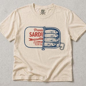 May include: A beige short-sleeve t-shirt featuring a graphic of an open sardine tin. The design includes blue and red ink with the text Premia SARDINES PACKED IN OLIVE OIL. The shirt has a crew neckline and a Comfort Colors brand label at the neck.