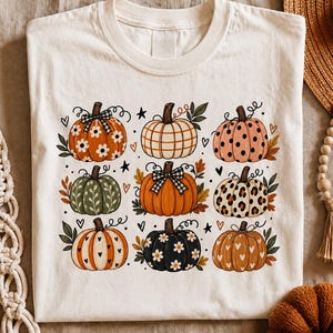Peut inclure: Un t-shirt couleur crème arborant une impression graphique de neuf citrouilles distinctes disposées en grille. Les citrouilles présentent divers motifs dont des marguerites, des carreaux, des pois, des taches de léopard et des cœurs.