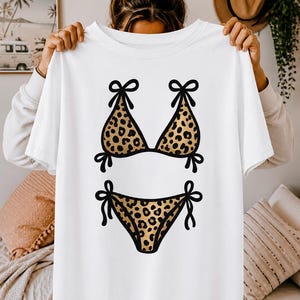 Peut inclure: T-shirt blanc avec un motif de bikini léopard. Le haut et le bas du bikini sont soulignés en noir et présentent un motif tacheté marron et beige. Le bikini a des détails de nœuds noirs.