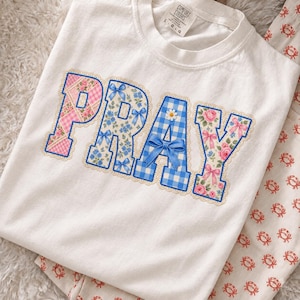 Könnte beinhalten: Ein weißes T-Shirt mit dem Wort PRAY in großen Applikationsbuchstaben. Jeder Buchstabe hat ein einzigartiges Muster: rosa Karo mit Rosen, blaue Blumen mit Schleifen, blaues Gingham mit Gänseblümchen und Schleife, sowie cremefarben mit Rosen und rosa Bändern.