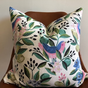 May include: Decorative throw pillow with a white background and a colorful bird and botanical pattern. The design features blue and pink birds, green leaves, and pink flowers. The pillow is on a brown chair.