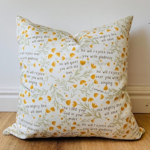 May include: A square decorative pillow with a floral pattern in yellow and green on a white background. The pillow features inspirational text from Zephaniah 3:17, in a handwritten style font.
