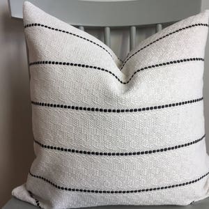 May include: A square, textured white pillow with horizontal black dotted stripes. The pillow is made of woven fabric and is placed on a gray chair. The design is simple and modern, suitable for home decor.