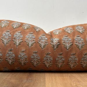 May include: A long, rectangular pillow with a burnt orange background and a repeating pattern of stylized, silver-colored floral motifs. The pillow is made of a soft, textured fabric and is placed on a wooden surface.