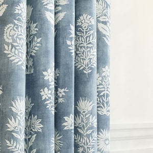 Op de afbeelding: Blauw en wit bloemmotief gordijn. De stof heeft een textuur met een vervaagde blauwe achtergrond. Witte bloemen- en bladmotieven zijn op het gordijn gedrukt. Het gordijn hangt in verticale plooien.