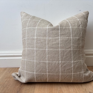 May include: A square decorative pillow with a neutral beige fabric and a white grid pattern. The pillow is placed on a wooden floor and leans against a white wall.