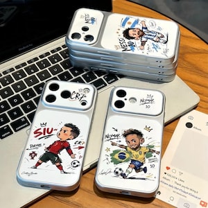 Könnte beinhalten: Drei silberfarbene Smartphone-Hüllen mit Cartoon-Illustrationen von Fußballspielern. Eine Hülle zeigt Messi im Argentinien-Trikot, eine weitere Ronaldo im Portugal-Trikot mit dem Text SIU und die dritte Neymar im Brasilien-Trikot.
