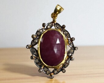 May include: Oval-shaped ruby pendant with a gold bail. The ruby is set in a gold frame, surrounded by a decorative silver-toned border. The pendant is displayed on a light-colored wooden surface.