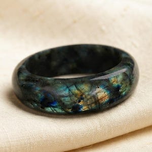 Könnte beinhalten: Ein polierter Bandring aus Labradorit-Stein mit einer dunkelgrauen Grundfarbe und irisierenden Blitzen in Blau, Grün und Gold auf der gesamten Oberfläche. Der Ring hat ein glattes, abgerundetes Profil und eine glänzende, reflektierende Oberfläche.